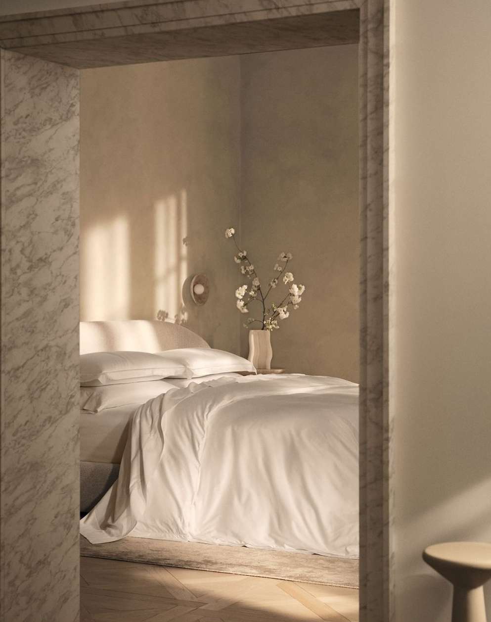 Bedroom interior featuring a bed with white linens, white cotton flowers in a vase, and light walls, viewed through a marble doorway.