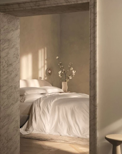 Bedroom interior featuring a bed with white linens, white cotton flowers in a vase, and light walls, viewed through a marble doorway.