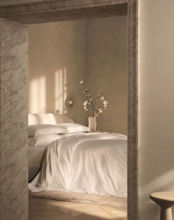 Bedroom interior featuring a bed with white linens, white cotton flowers in a vase, and light walls, viewed through a marble doorway.