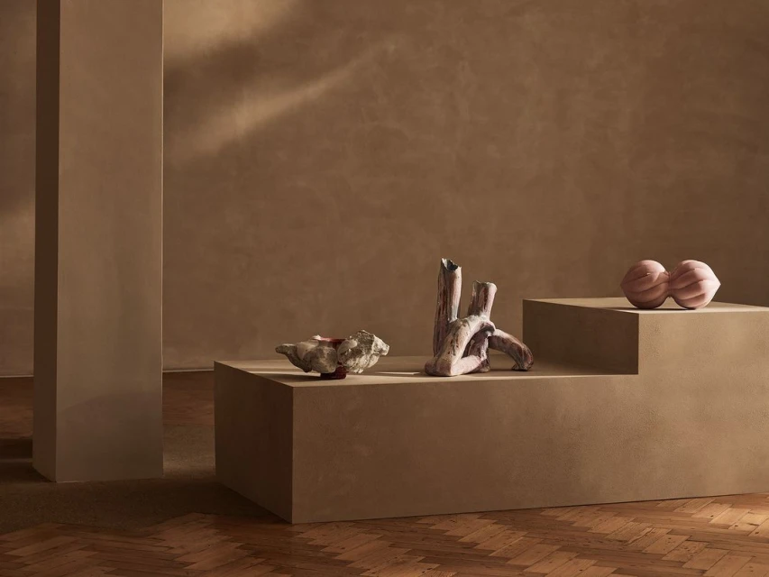 Abstract interior shot featuring geometric art pieces on podiums against a textured brown wall, with a herringbone wood floor.