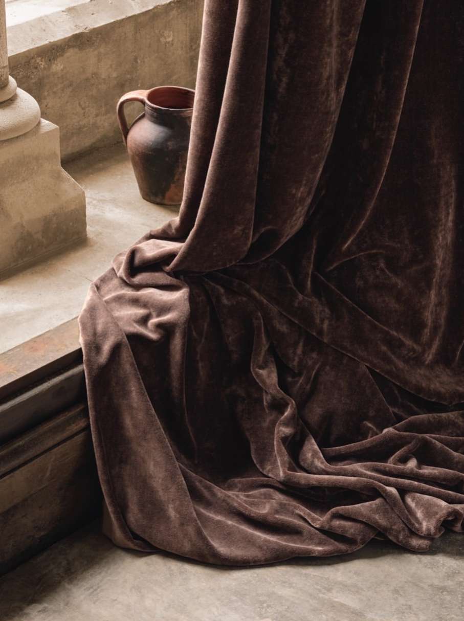 Still life featuring a brown ceramic jug and draped brown velvet fabric with soft lighting against a textured, neutral-toned wall.