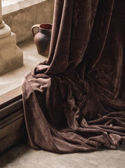 Still life featuring a brown ceramic jug and draped brown velvet fabric with soft lighting against a textured, neutral-toned wall.