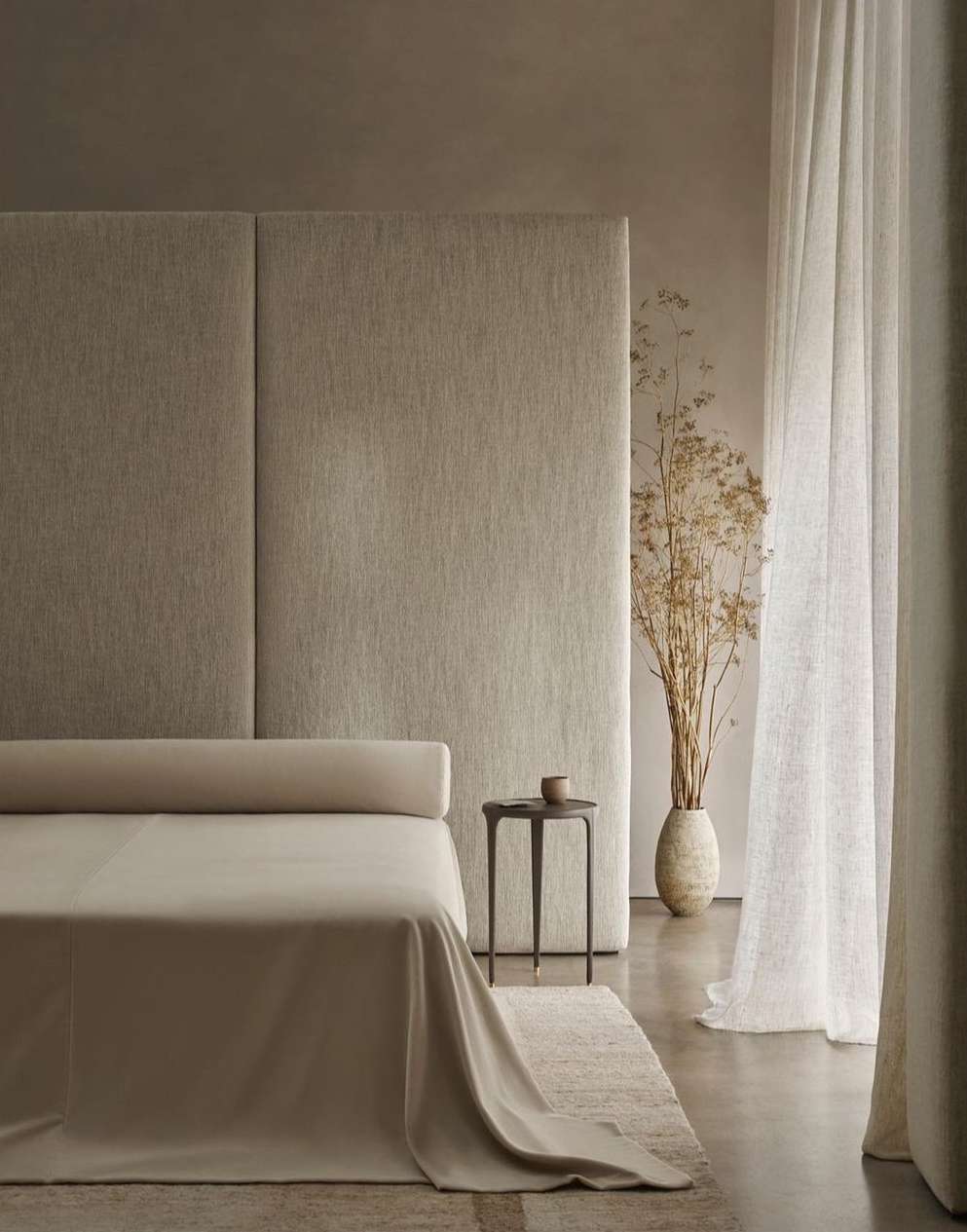 Beige bedroom interior with linen bedding, a dark side table, a tall vase with dried flowers, and sheer white curtains, creating a neutral and calming space.