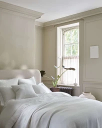 A tranquil white bed in a neutral paneled bedroom with natural light streaming through a traditional arched window.