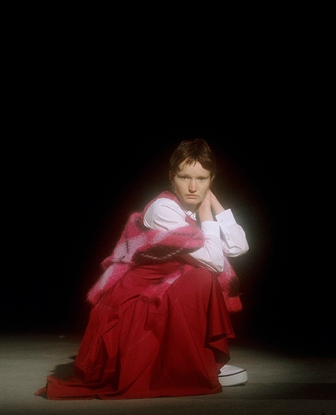 Woman in red skirt and white shirt sits in front of a black background, lit by spotlight. She wears a colorful shawl.