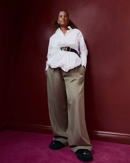 An adult female standing in a photo studio with a white shirt, wide black belt, and tan trousers against a red wall and fuchsia carpet.