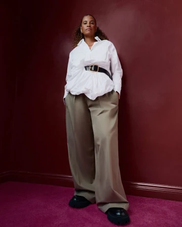 An adult female standing in a photo studio with a white shirt, wide black belt, and tan trousers against a red wall and fuchsia carpet.