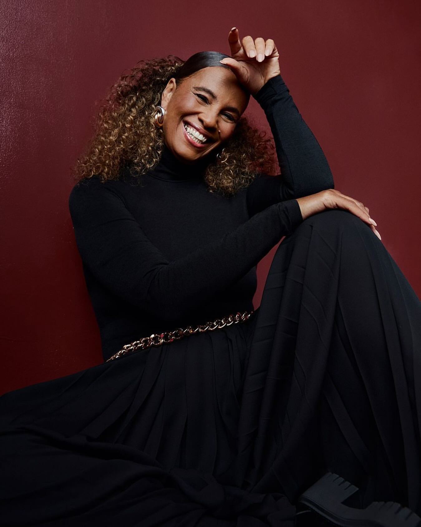 A smiling Black woman in a long black dress and a gold chain belt, seated against a dark red backdrop, exudes confidence and style.