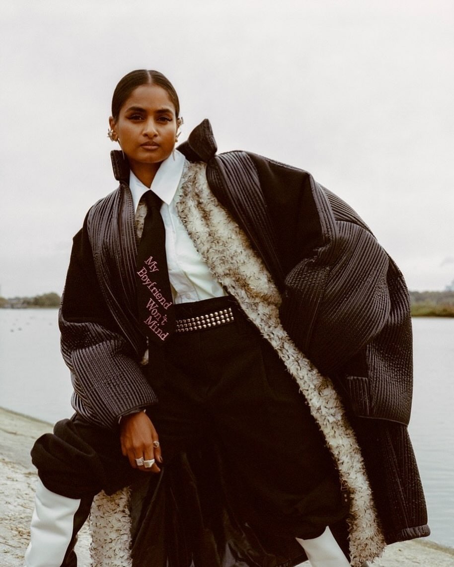 Fashion editorial shot of a South Asian woman wearing a puffy coat, tie, and studded belt outdoors with water and sky in the background.