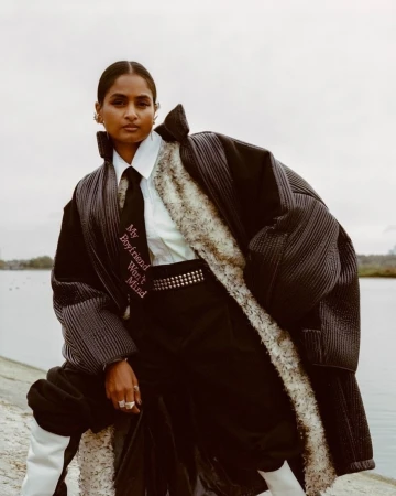 Fashion editorial shot of a South Asian woman wearing a puffy coat, tie, and studded belt outdoors with water and sky in the background.