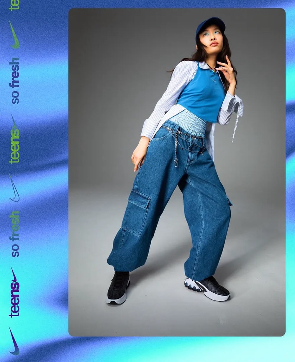 Stylish young woman in oversized jeans, blue top, and cap poses in a studio with blurred blue background and Nike logo branding.
