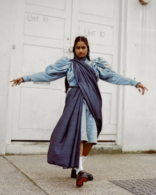 Woman in blue dress with draped fabric poses in front of white door with graffiti. Fashion editorial style with urban backdrop.