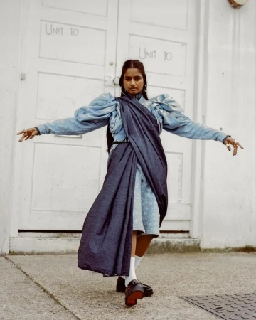 Woman in blue dress with draped fabric poses in front of white door with graffiti. Fashion editorial style with urban backdrop.
