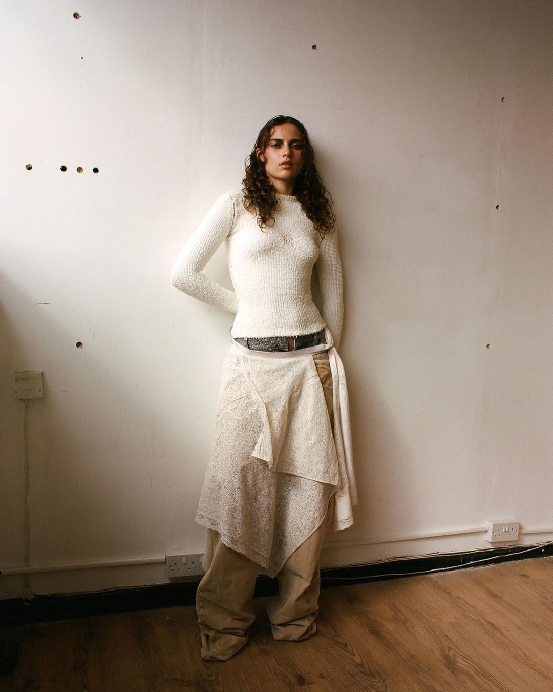 A woman in an ivory sweater and layered skirt poses indoors against a white wall with a wood floor, showcasing a fashion style.