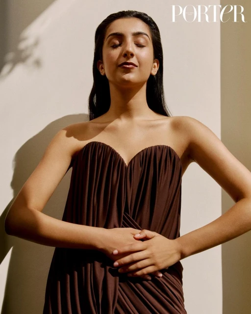 A young woman poses in a draped brown strapless dress with her eyes closed, bathed in sunlight with 'PORTER' text in the corner.
