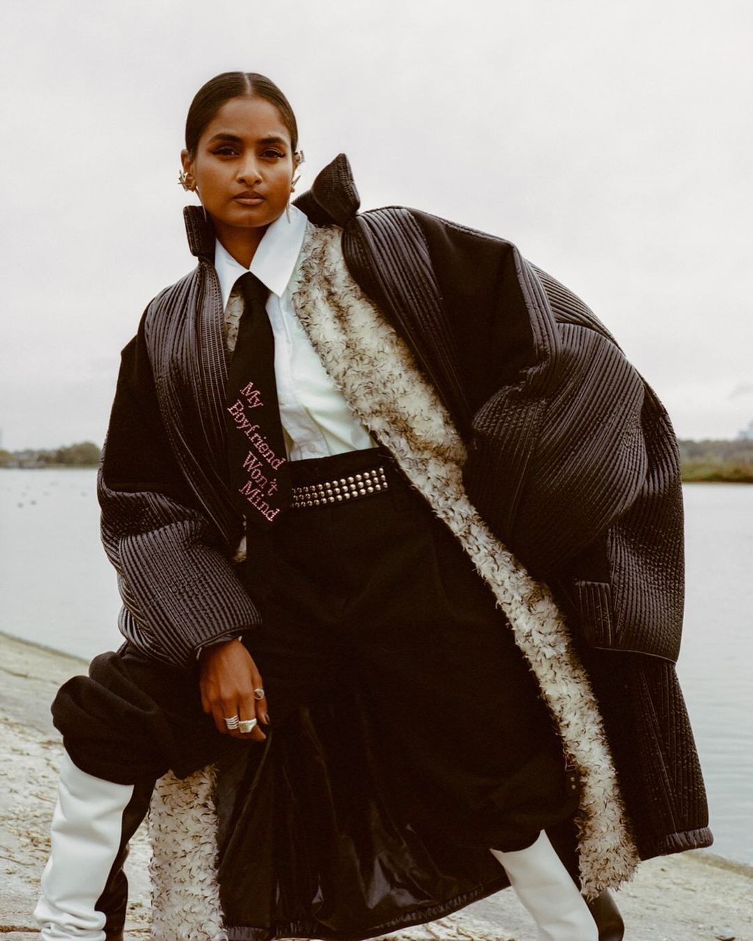 Woman in oversized black coat with tie poses outdoors. Moody editorial fashion portrait with overcast sky. Beauty shot with fashion clothing.