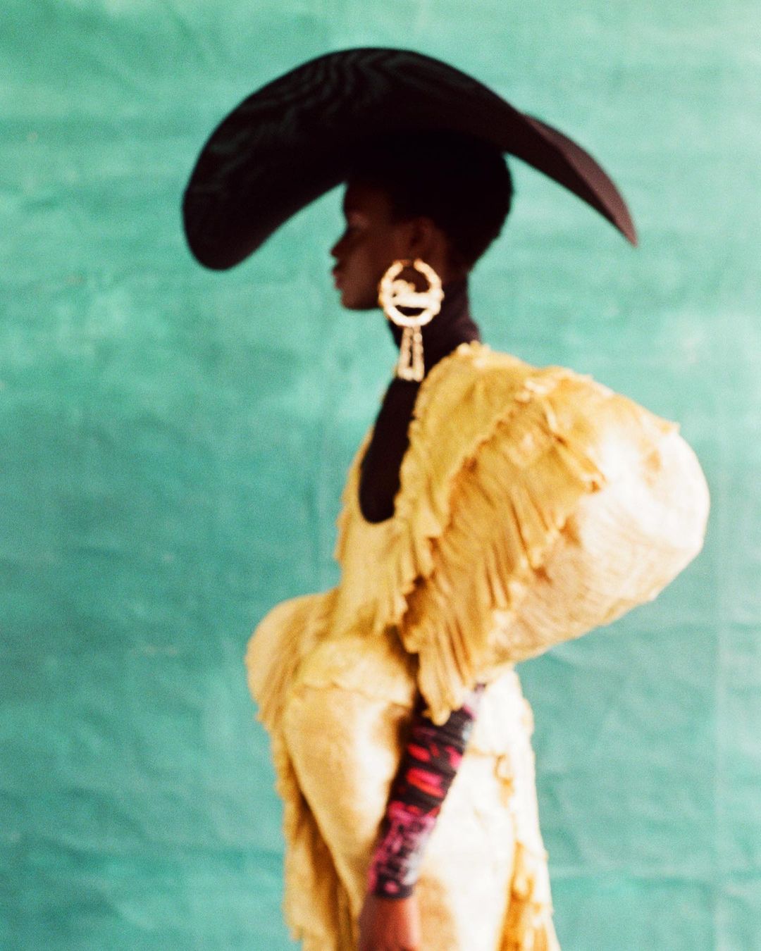 A stylish black woman in profile, wearing a large black hat and a gold dress with dramatic sleeves, posing in front of a mottled turquoise background.