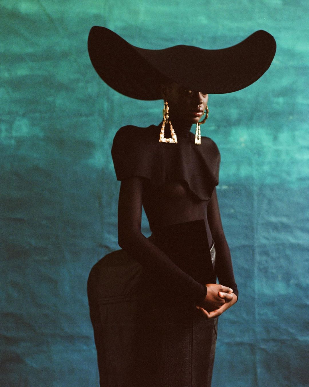 Portrait of an adult woman in a large black hat, black top and skirt, and dangling triangle earrings against a blue mottled backdrop.