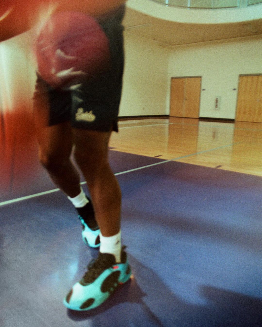 A person dribbles a basketball on a hardwood court inside a gymnasium, wearing shorts and athletic shoes, with light streaming in.