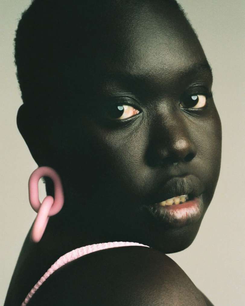 Close-up portrait of a dark-skinned adult female with short dark hair, a pink tank top, and pink hoop earrings against a gray background.
