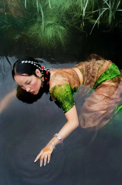 An Asian woman in a translucent dress kneels in dark water, with green plants in the background, creating a dreamy, ethereal atmosphere.