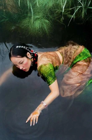 An Asian woman in a translucent dress kneels in dark water, with green plants in the background, creating a dreamy, ethereal atmosphere.