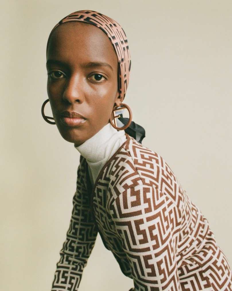 Portrait of an adult female wearing a patterned dress and headscarf, along with hoop earrings, against a light background in a studio setting.