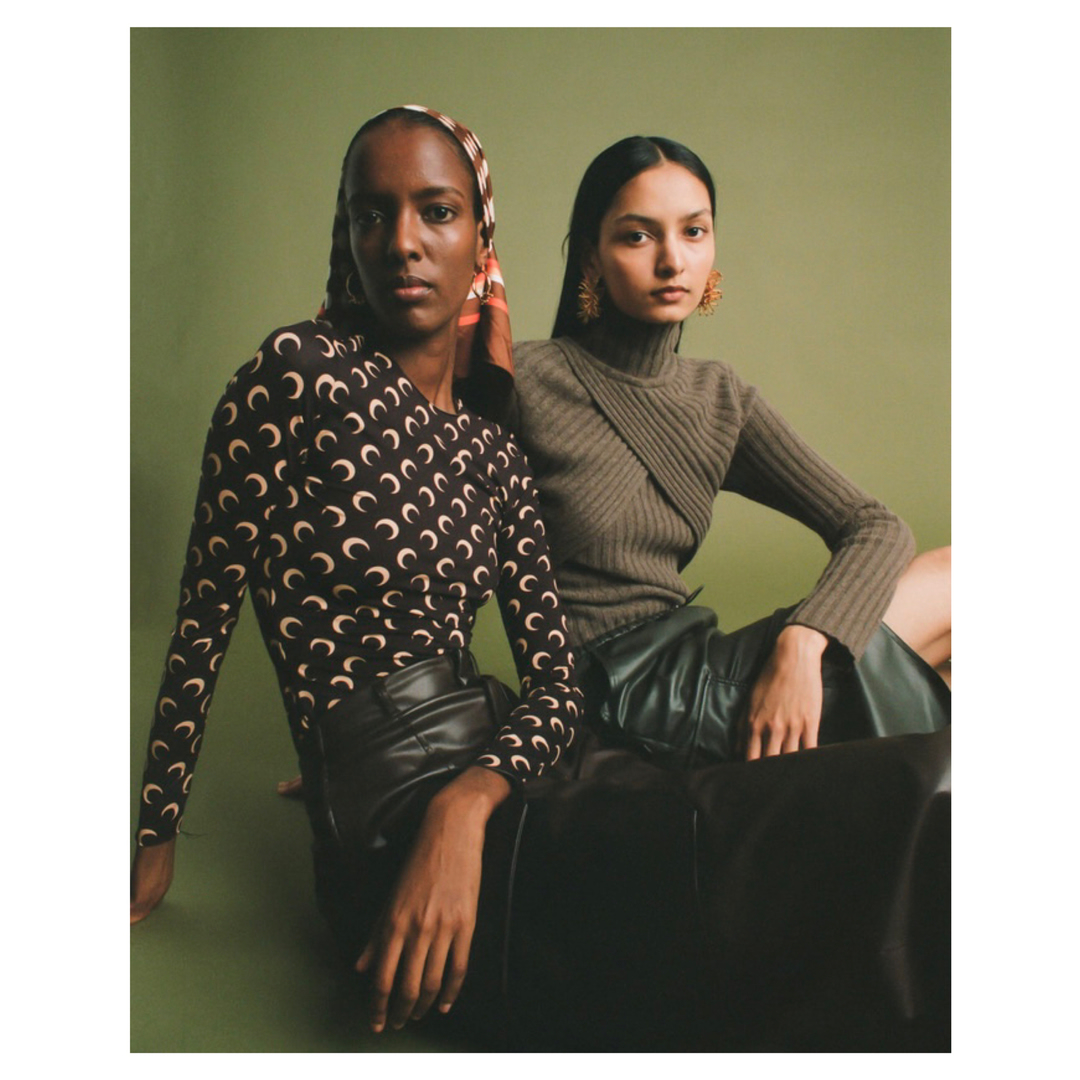 Two women in stylish tops and skirts pose in a studio, one in a crescent moon top and headscarf, the other in a turtle neck sweater.
