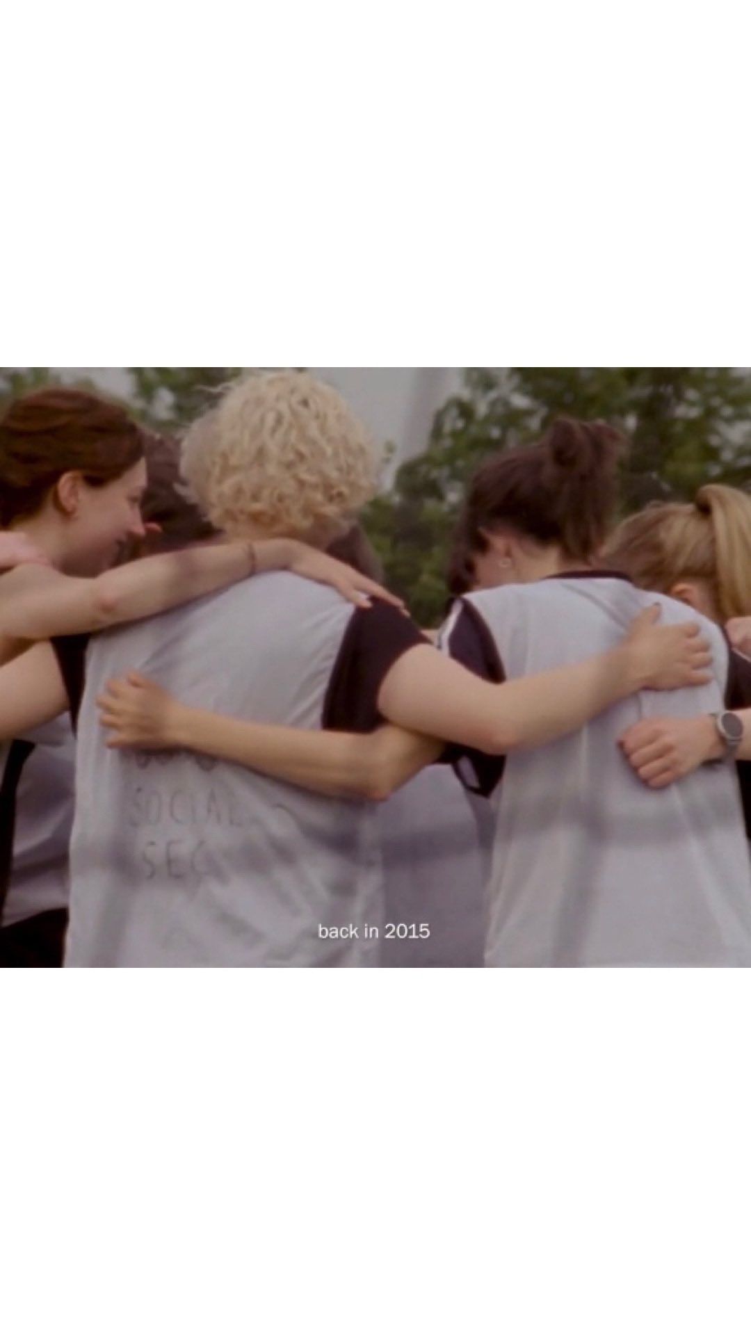 Group of young women embracing, backs to camera, wearing similar shirts, in an outdoor setting, close together, with a sense of camaraderie.