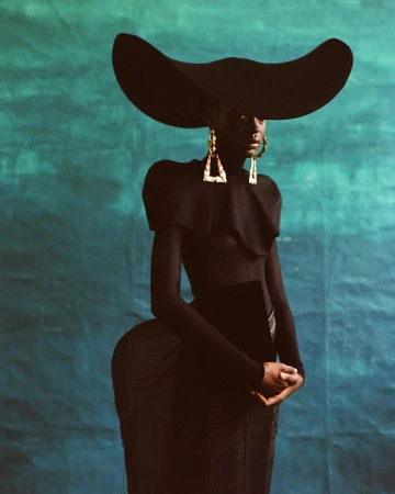 Model in large wide brim black hat and geometric gold earrings wearing a black top against a textured teal backdrop
