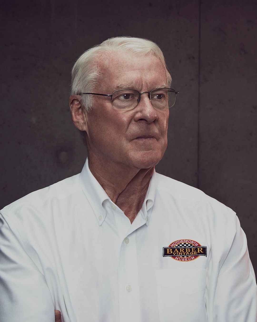 Portrait of an elderly adult man wearing glasses and a white collared shirt with a logo patch, against a grey textured background.