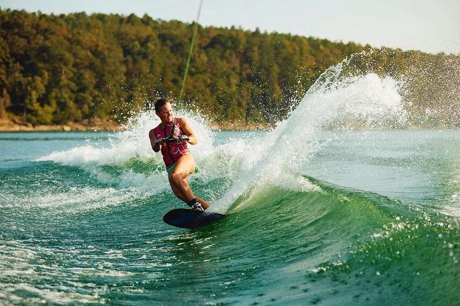 A woman wakeboarding on a lake, captured in a dynamic action shot with splashing water and a backdrop of forested landscape.