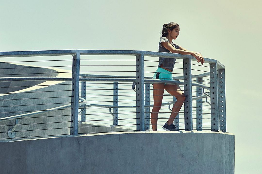 Young woman in athletic wear stands on an urban balcony overlooking a bright skyline on a sunny day, shot in medium framing.
