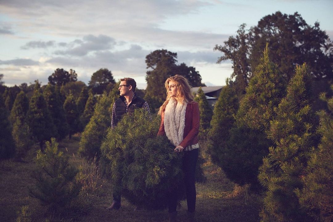 A young couple carries a fresh-cut Christmas tree through a tree farm at dusk, with rows of evergreens and a cloudy sky.