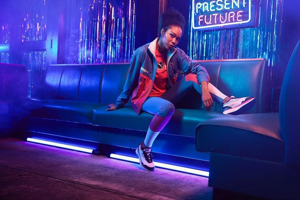 An adult female model sits on a blue sofa wearing a Puma jacket and leggings in a neon-lit indoor environment with reflective tinsel.