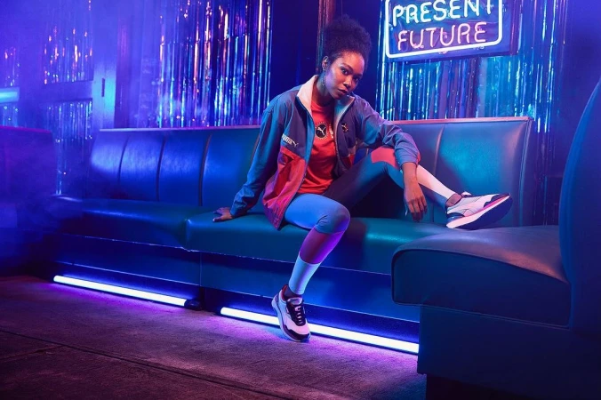 An adult female model sits on a blue sofa wearing a Puma jacket and leggings in a neon-lit indoor environment with reflective tinsel.