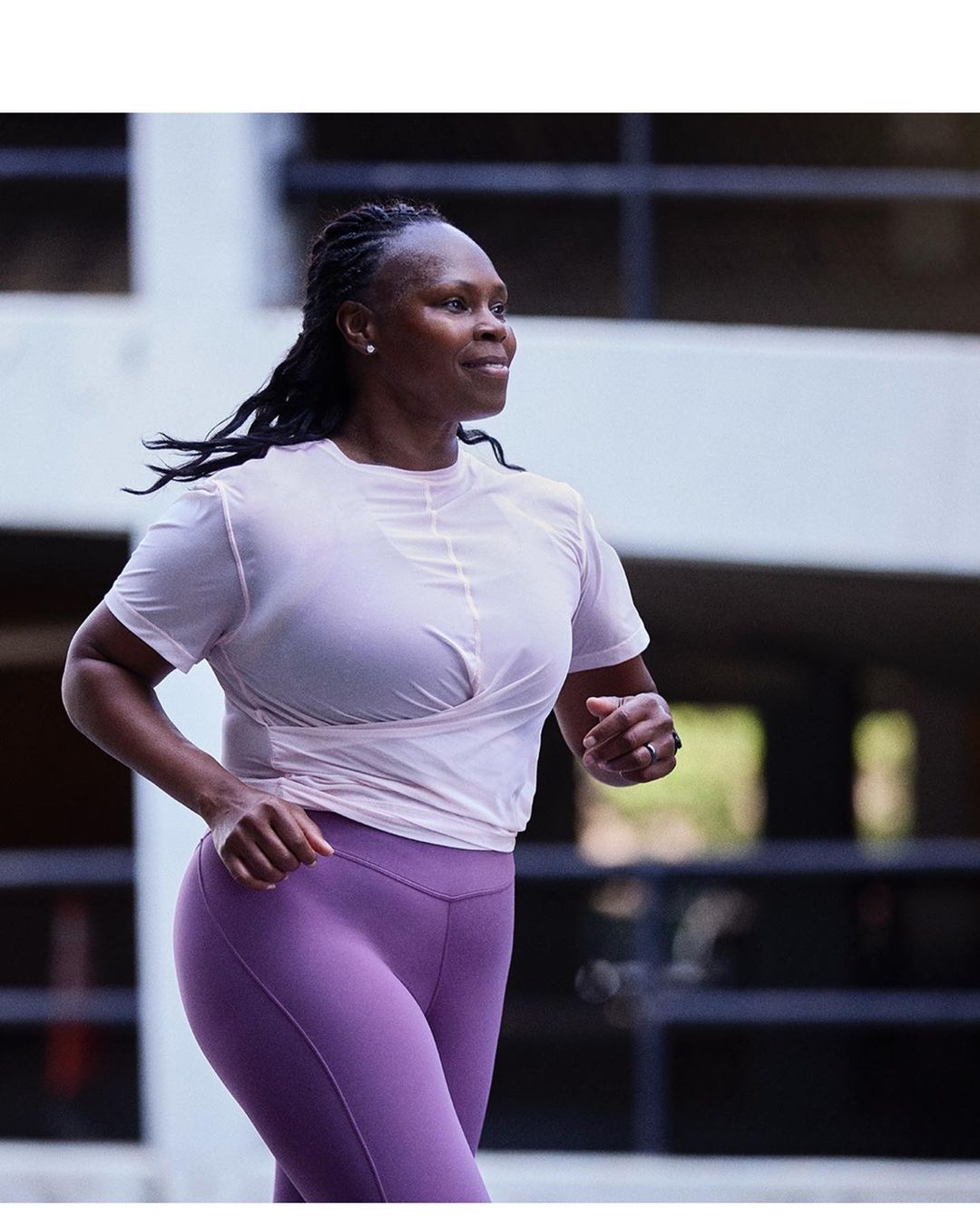 Athletic woman with braided hair running outdoors in a light pink athletic shirt and purple leggings in an urban environment.