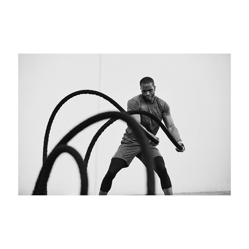 An African-American man is working out with battle ropes indoors, wearing athletic shorts and knee pads, captured in a black and white photograph.