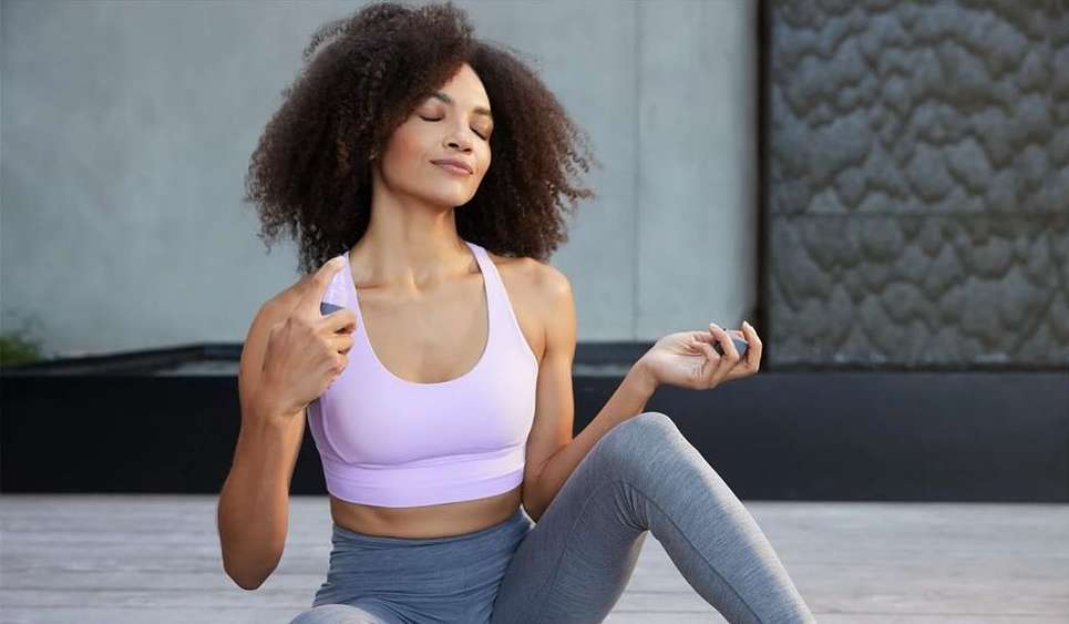 A woman with curly hair sits cross-legged wearing a lavender sports bra and grey leggings, meditating with her eyes closed outdoors.