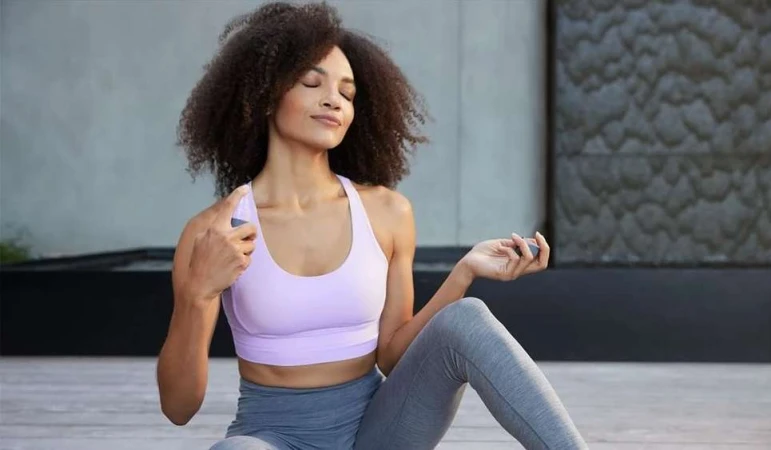 A woman with curly hair sits cross-legged wearing a lavender sports bra and grey leggings, meditating with her eyes closed outdoors.