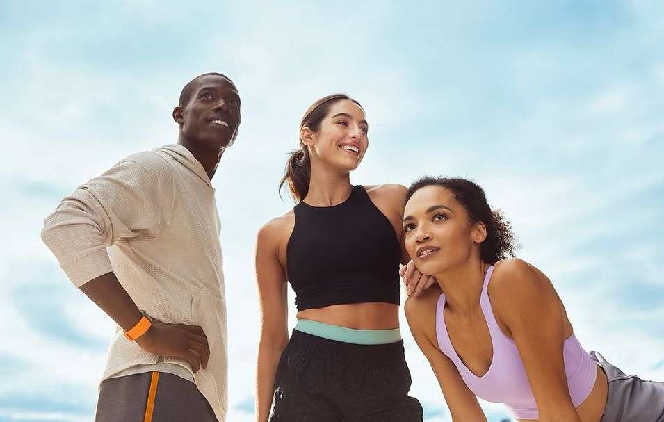Three smiling, diverse people stand outdoors in activewear against a bright sky; one man, two women; fitness and healthy lifestyle promotion.