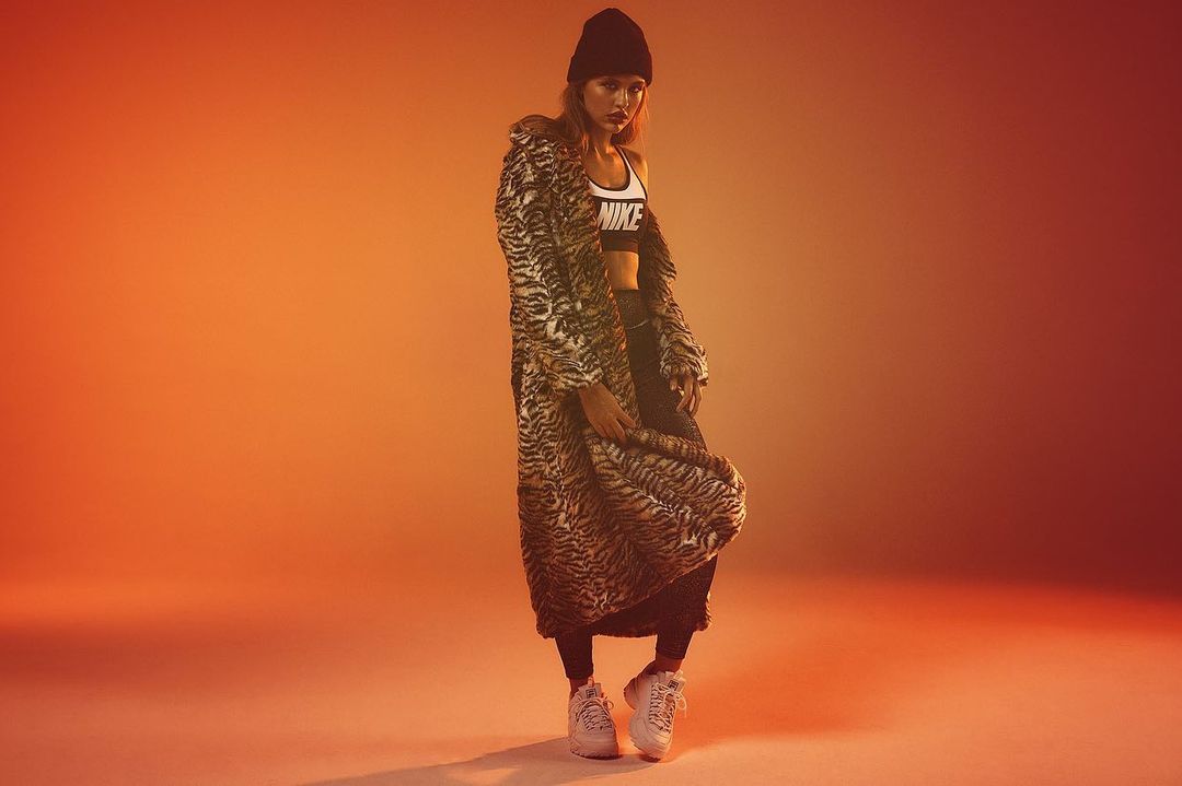 Woman in a tiger-striped coat poses confidently in a studio wearing a black beanie, bra top with the Nike logo, and sneakers on a warm gradient backdrop.
