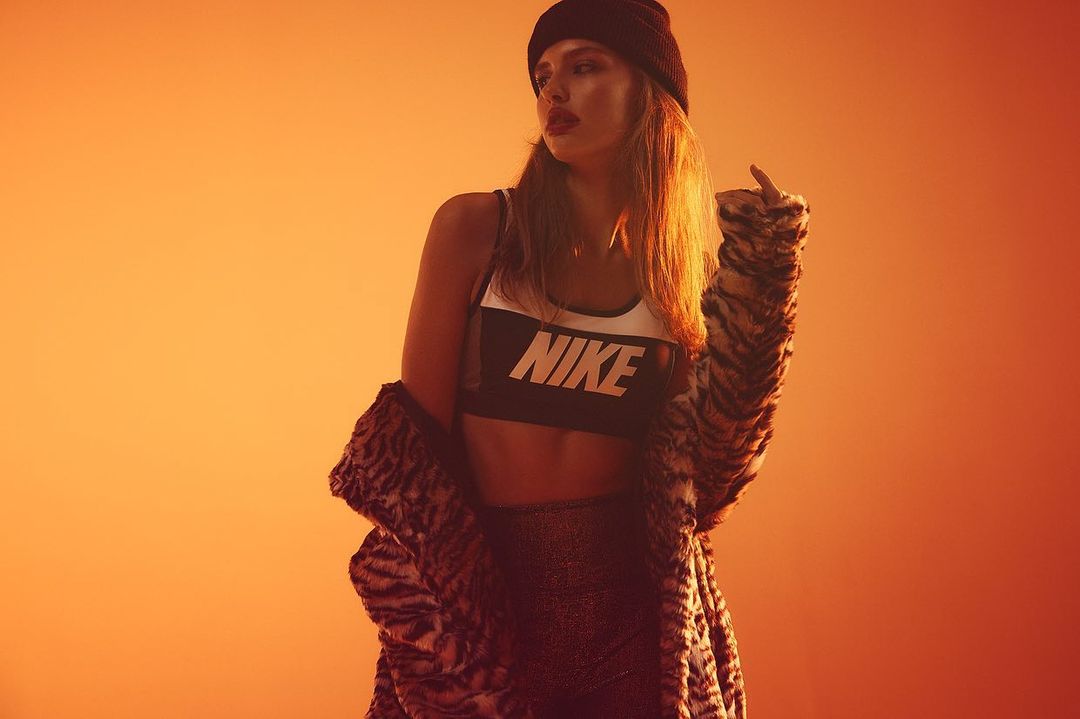 A young woman poses against an orange backdrop, wearing a black beanie, athletic bra with 'NIKE' logo, and a patterned faux fur coat.