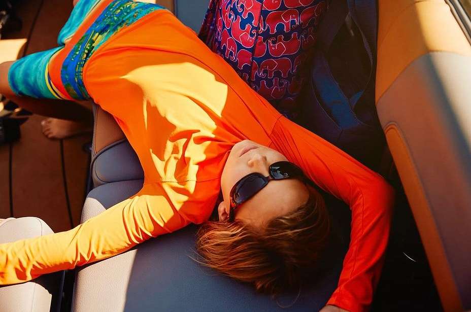 Young adult rests on a boat wearing sunglasses and bright orange long sleeve shirt, enjoying sunny weather and leisure on the water.