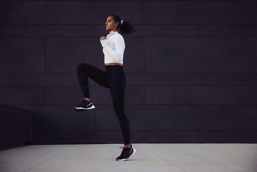 Athletic woman in fitness attire performs high knees exercise outdoors against a dark grey textured wall, promoting health and wellness.