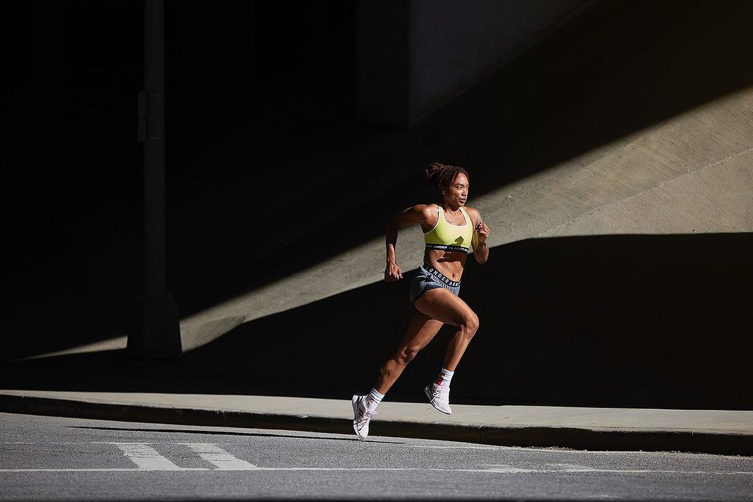Athletic Black woman in workout clothes powerfully running outdoors under an underpass. She is training in sunlight with a determined expression.