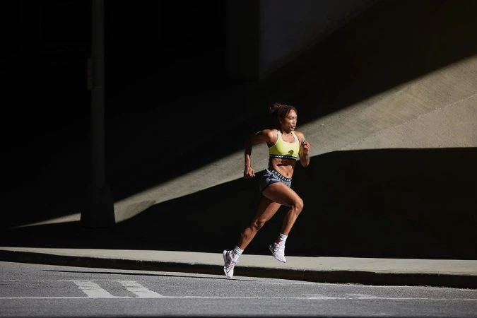 Athletic Black woman in workout clothes powerfully running outdoors under an underpass. She is training in sunlight with a determined expression.