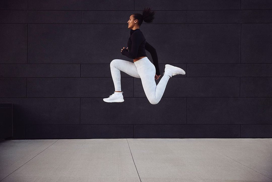Athletic woman in black top and patterned white leggings mid-air in dynamic leap against a dark tiled wall in an urban setting.
