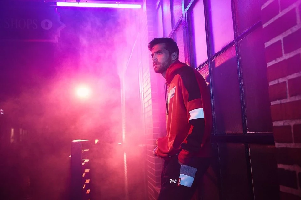 Man in a red hoodie poses in an urban setting with pink and purple lighting, brick wall, and doorway, looking away from the camera.