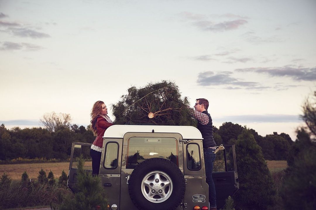A man and woman load a Christmas tree onto the roof of a gray Land Rover against a backdrop of a wintery landscape with blue skies.